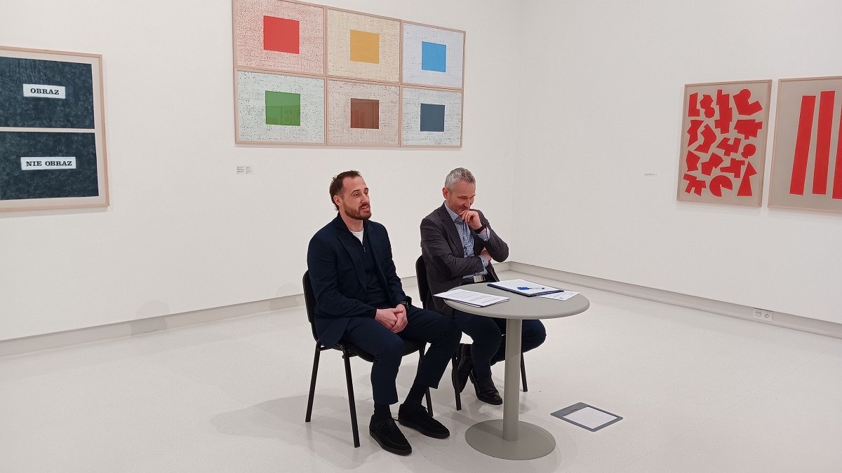 dwóch mężczyzn siedzi przy stoliku w białej sali, na jej ścianach wiszą obrazy przedstawiające sztukę nowoczesną