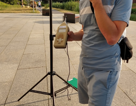 osoba stojąca i trzymająca urządzenie do pomiaru hałasu, nie widać jej twrazy w kadrze - grafika artykułu
