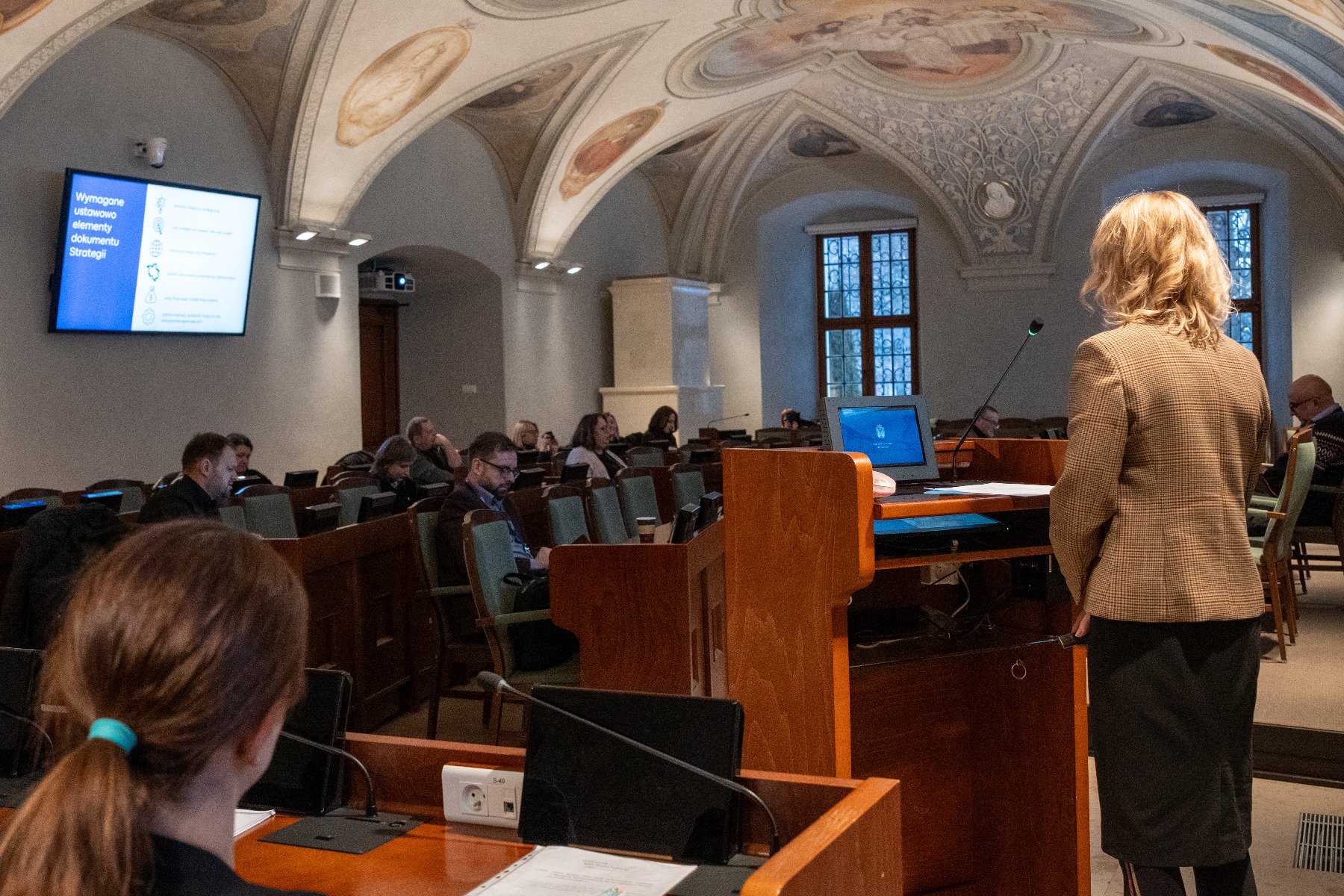 Sala sesyjna. Osoba mówiąca stoi przy mównicy, uczestnicy siedzą w lożach w tle.