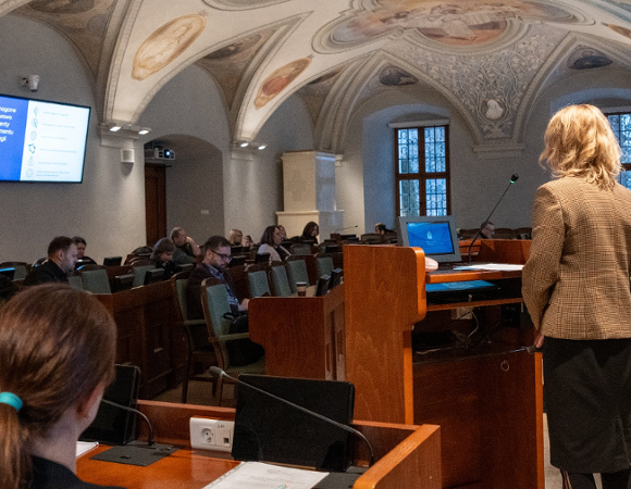 Sala sesyjna. Osoba mówiąca stoi przy mównicy, uczestnicy siedzą w lożach w tle.