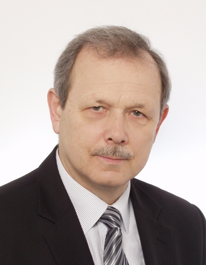 Mężczyzna w średnim wieku, częściowo siwiejące włosy i wąs, ubrany w ciemną marynarkę i jansą koszulę, delikatnie się uśmiecha