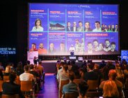 w dużej sali konferencyjnej wiele osób siedzi na krzesłach ustawionych w rzędach, sala jest przyciemniona, na dużym ekranie wyświetlana jest prezentacja