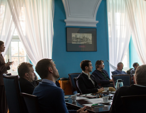 w sali konferencyjnej przy stole siedzi kilka osób, inne osoby siedzą na krzesłach za stołem, jedna osoba stoi i przedstawia prezentację
