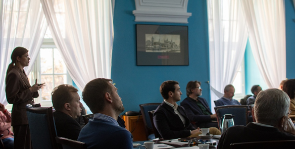 w sali konferencyjnej przy stole siedzi kilka osób, inne osoby siedzą na krzesłach za stołem, jedna osoba stoi i przedstawia prezentację