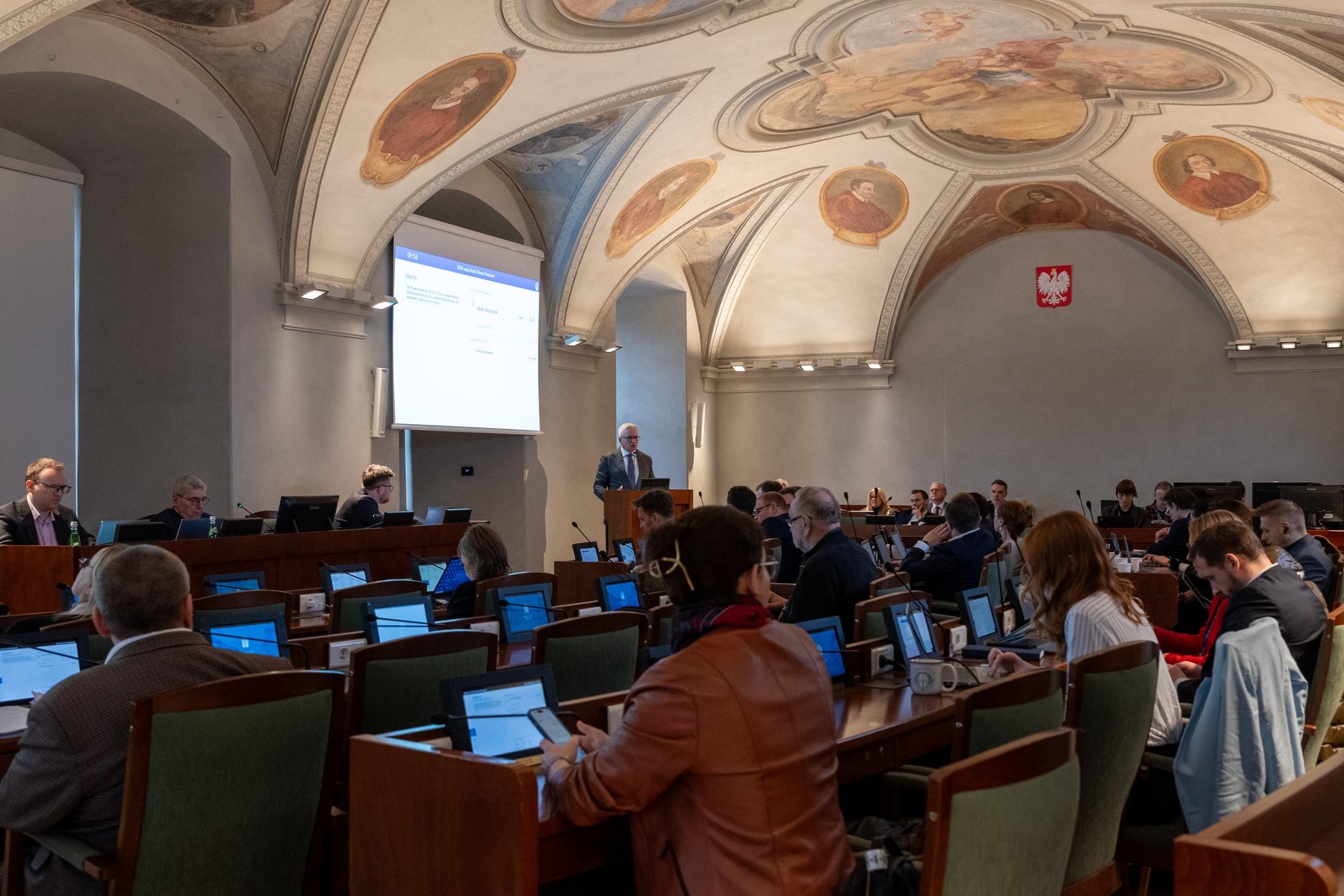 Zdjęcie przedstawia przebieg rady miasta. Radni siedzą przy lożach.
