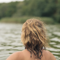 Kobieta w wodzie, widać plecy i długie włosy. Z przodu las.