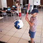 Sala, stoliki i krzesła. Na stołach laptopy. N apierwzym planie chłopiec z akrytymi czapka oczami z piankowa rurka próbuje trafić w zawieszona piłkę. Dalej 3 dzieci przy stolikach.