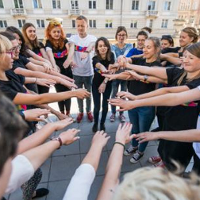 Młodzież stoi na placu w kręgu z wyciągniętymi rękoma do środka. W tle budynki.