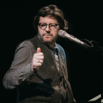 Na zdjęciu artysta siedzący przy fortepianie z mikrofonem przy twarzy.
