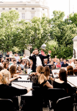Photo of a orchestra performing outside. The conductor in front of the orchestra, the audience behind him, green trees as a background.
