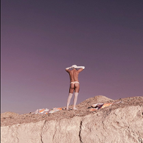 A picture of half-naked man standing on a rock, wearing white knee socks, long white gloves, white cap and white thong.