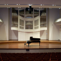 Zdjęcie przedstawia scenę auli Akademii Muzycznej w Poznaniu. Na środku sceny ustawiony jest fortepian. W tle widoczne organy.