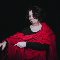 Kobieta w czarno-czerwonym stroju stoi na ciemnym tle.