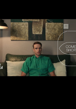 Kadr obrazu na którym widzimy mężczyznę w zielonej koszuli z krótkim rękawem, siedzącego na zielonej dwuosobowej kanapie z dwoma białymi poduszkami za plecami. W tle również lampa stojąca, zaświecona oraz fragment obrazu nad kanapą. Na kadr nałożono logo festiwalu oraz cyklu comedy shorts.