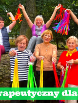 Grupa uśmiechniętych seniorek w kolorowych ubraniach. Machają kolorowymi wstążkami. W tle pień drzewa i zieleń. Tytuł wydarzenia na dole, na zielonym tle.