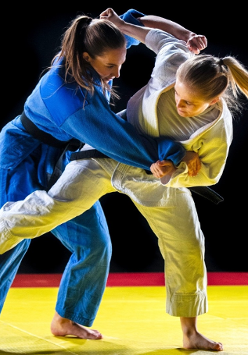 Dwie zawodniczki judo ćwiczące na macie. Jedna ubrana w białą a druga w niebieską judogę