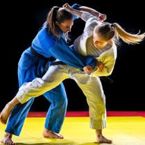 Dwie zawodniczki judo ćwiczące na macie. Jedna ubrana w białą a druga w niebieską judogę