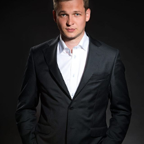 Na zdjęciu widoczny jest mężczyzna ubrany w elegancki czarny garnitur i białą koszulę. Stoi przed ciemnym tłem, które podkreśla jego sylwetkę. Mężczyzna ma krótkie jasne włosy i patrzy bezpośrednio w kamerę z lekkim uśmiechem. Ogólny ton fotografii tworzy wrażenie profesjonalizmu i pewności siebie.