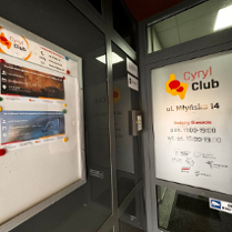 Zdjęcie przedstawia wejście do klubu o nazwie "Cyryl Club" w Poznaniu, Polska. Klub znajduje się na ulicy Młyńskiej, numer 14. Na drzwiach znajduje się tabliczka z nazwą i adresem klubu, a także godzinami otwarcia. Po lewej stronie od wejścia znajduje się tablica z plakatami reklamującymi nadchodzące wydarzenia, a także czerwone i niebieskie magnesy. Dodatkowo na drzwiach znajduje się naklejka informująca, że lokal jest monitorowany.