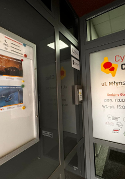 Zdjęcie przedstawia wejście do klubu o nazwie "Cyryl Club" w Poznaniu, Polska. Klub znajduje się na ulicy Młyńskiej, numer 14. Na drzwiach znajduje się tabliczka z nazwą i adresem klubu, a także godzinami otwarcia. Po lewej stronie od wejścia znajduje się tablica z plakatami reklamującymi nadchodzące wydarzenia, a także czerwone i niebieskie magnesy. Dodatkowo na drzwiach znajduje się naklejka informująca, że lokal jest monitorowany.