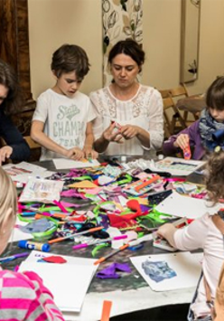 Dzieci z opiekunami na zajęciach przy stole pełnym kolorowych papierów.