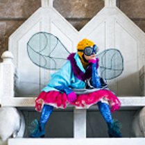 Kobieta przebrana w kolorowe ciuchy za muchę siedząca na białej ławie.