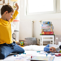Prawdopodobnie ojciec z synem grają na podłodze w plamszówkę. Chłopiec cieszy się, ma podniesione rece do góry. Na stołeczku leży kilka gier planszowych.