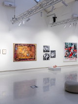 Sala wystawowa z obrazami zawieszonymi na ścianach, pod sufitem metalowa konstrukcja z oświetleniem, pośrodku podłogi pufa.