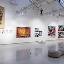 Sala wystawowa z obrazami zawieszonymi na ścianach, pod sufitem metalowa konstrukcja z oświetleniem, pośrodku podłogi pufa.