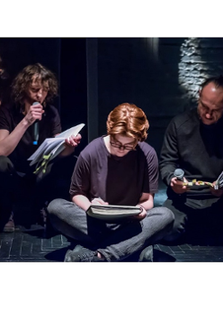 Kilku mężczyzn siedzi na scenie, każdy z nich trzyma przed sobą otwartą książkę. Jeden z mężczyzn patrząc na otwartą książkę czyta do mikrofonu.