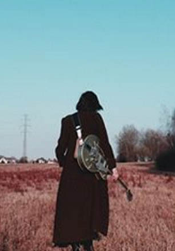 Foto. Kobieta z gitarą idzie przez pole.