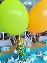 Balony na uchwytach w wysokiej szklance, na stole, obok dekoracje z papieru.