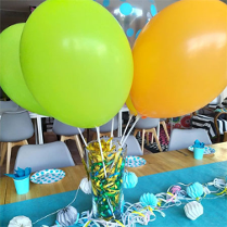 Balony na uchwytach w wysokiej szklance, na stole, obok dekoracje z papieru.