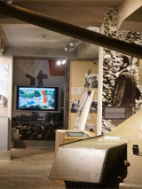 Ekspozycja w muzeum: po lewej na scianie monitor z wyswietlona postacią, w dłebi duży monitor, po prawej czołg i duże zdjęcia na scianie postaci.