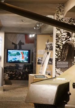 Ekspozycja w muzeum: po lewej na scianie monitor z wyswietlona postacią, w dłebi duży monitor, po prawej czołg i duże zdjęcia na scianie postaci.