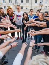 Młodzież stoi na placu w kręgu z wyciągniętymi rękoma do środka. W tle budynki.