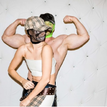 Męszczcyzna pręży muskuły. Przed nim kobieta. Oboje w czapkach z daszkiem z zasłonietymi twarzami. Kobieta ma zasłoniete bandażem piersi.