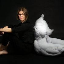 Krystyna Lipka-Czajkowska siedzi na podłodze, ręce położyła na kolanach, oparta o uszytą rzeźbę kobiety ułożonej w podobnej pozycji.