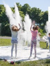 Fotografia przedstawia dzieci rzucające białym proszkiem ponad białym materiałem rozłożonym na trawie. Dodatkowo na fotografię nałożono logo 21. edycji Letniego Pogotowia Sztuki.