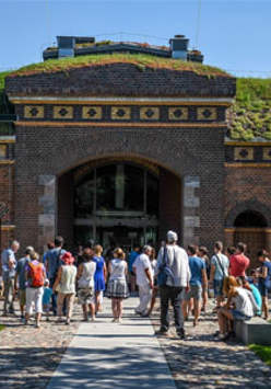 Budynek śluzy z czerwonej cegły. Na dachu trawa. Przed wejściem najprawdopodobniej wycieczka.