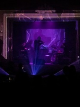 Ciemna fotografia została wykonana na sali koncertowej. W jej centrum widzimy niewyraźnie sylwetkę skrzypka stojącego i grającego na scenie, a za nim sylwetki kilku innych muzyków. Zdjęcie zrobiono ponad głowami osób siedzących na widowni. Utrzymane jest fioletowych barwach światła padającego spod sceny.