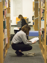 Fotografia dwóch kobiet kucających między regałami z książkami i czytającymi książki.