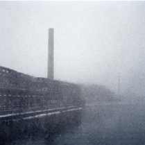 Stara fotografia przedstawiająca budynek z kominem stojący na brzegu rzeki.