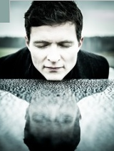 Portret muzyka w zbliżeniu. Jego głowa spoczywa na fortepianie, na powierzchni dostrzec można jej lustrzane odbicie. W tle otwarty leśny krajobraz.