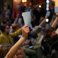 Fotografia: publiczność slamu sfotografowana podczas głosowania. Zdjęcie zostało zrobione nocą, jest nieostre. Na pierwszym planie podniesiona do góry ręka z białą kartką, w tle inni widzowie, którzy także głosują w ten sam sposób, podnosząc rękę do góry.