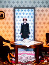 Na grafice kobieta i mężczyzna siedzący przy stole. W środku grafiki dziewczynka stojąca w drzwiach.