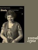 Na oliwkowym tle okładka książki Joanny Ostrowskiej pod tytułem "Ślady. Historia pewnego archiwum". Na okładce uśmiechnięta kobieta w krótkich włosach, ubrana w kwiecistą sukienkę i kamizelkę.