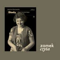 Na oliwkowym tle okładka książki Joanny Ostrowskiej pod tytułem "Ślady. Historia pewnego archiwum". Na okładce uśmiechnięta kobieta w krótkich włosach, ubrana w kwiecistą sukienkę i kamizelkę.