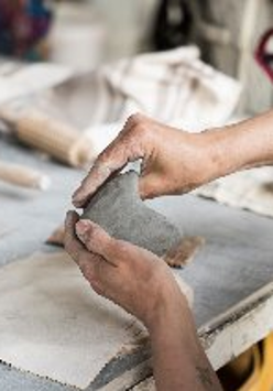 Kobieta modelująca ręcznie gliniany przedmiot na stole warsztatowym, otoczona narzędziami do ceramiki i materiałami do pracy.