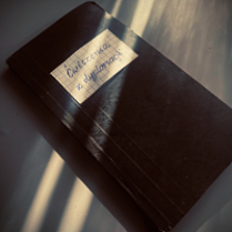 Na zdjeciu widoczny czarny zeszyt, na okładce w gornej części naklejony prostokąt z kartki zeszytowej w kratkę, z napisem "Ćwiczenia z dyplomacji".